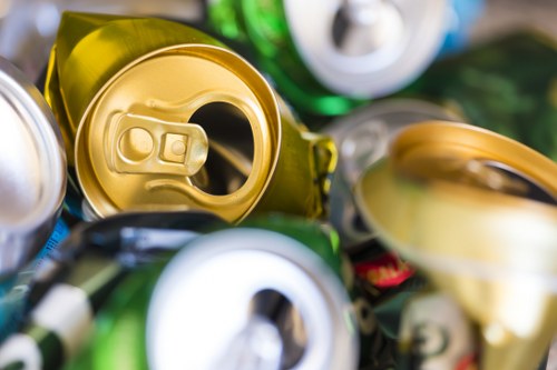 Close-up image of crushed aluminium beverage cans, predominantly featuring gold and silver colours with some hints of green from the can tops, piled together in a container on a pavement. The cans show creases, dents, and textures characteristic of used drink cans, with some slight reflections of light on their metallic surfaces. The background is blurred, suggesting an outdoor or urban setting, possibly on a driveway or service area in Shortlands, with part of a green plastic container or trash bin visible in the upper right corner. This image relates to waste collection and rubbish removal, and it visually represents recyclable materials that a commercial waste removal service such as Commercial Waste Removal Shortlands can handle, especially involving the collection of aluminium cans for disposal or recycling purposes.