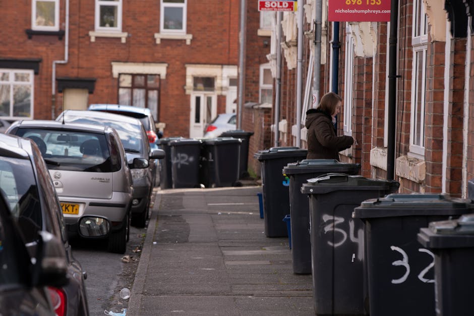 Five large outdoor rubbish bins lined up against a plain, light grey wall on a concrete surface. Each bin is made of dark grey plastic with a textured finish and features a bright yellow lid with a sl