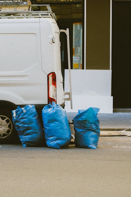 A white commercial van parked on a street with its rear doors closed, positioned next to a modern storefront with large glass windows and a dark frame. In front of the van, three large blue rubbish ba